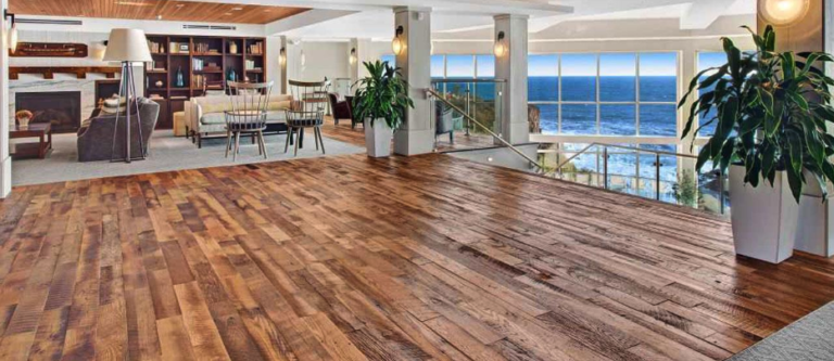Spacious open-plan living area with reclaimed wood flooring and modern furnishings. A dining table is set near a wall with bookshelves and a fireplace. Large windows offer an ocean view, and potted plants add greenery to the space, providing fresh design ideas for any home.