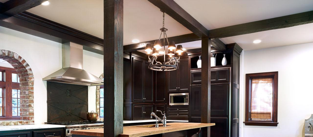 A modern kitchen featuring dark wood cabinetry, a central island with a reclaimed wooden countertop, a contemporary chandelier, and stainless steel appliances. Brick archways frame the space while natural light filters through a window with wooden blinds.