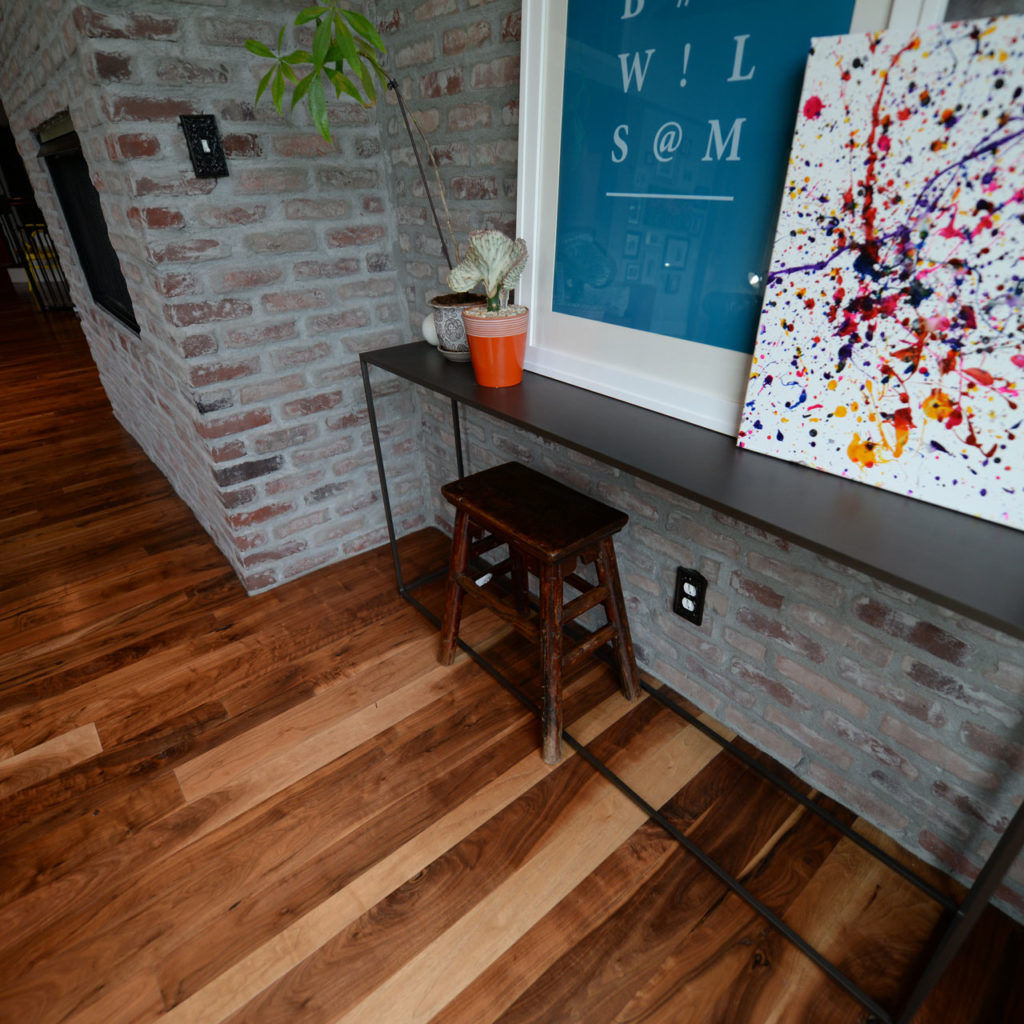 A rustic room with dark wood flooring and a brick wall exudes charm. A narrow table holds a small wooden stool, a potted plant, and an abstract painting with colorful splatters. Above the table, a framed blue poster showcases creative design ideas.