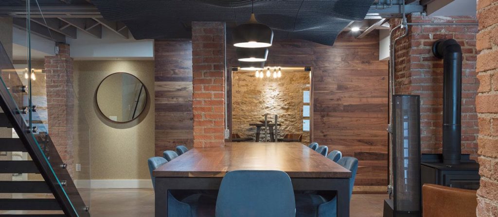 A modern dining area features a long wooden table surrounded by blue chairs. With exposed brick and stylish wall designs, the room exudes charm. Hanging pendant lights illuminate the space, enhancing its cozy atmosphere, while a staircase on the left adds to the allure.