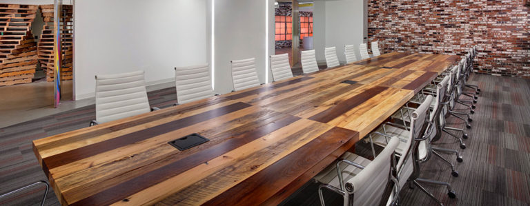 A long wooden conference table crafted from reclaimed wood is surrounded by white chairs in a modern office space. One brick wall contrasts with the carpeted floor, and the table tops come equipped with built-in power outlets. Dividers are partially visible in the background, blending style and function.