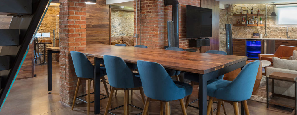 A modern dining area features a wooden table with sleek table tops and eight teal chairs in an industrial-style room. Exposed brick and stone walls, a mounted TV, shelves, and a bar area with blue lighting enhance the contemporary aesthetic, perfect for both residential and commercial design.