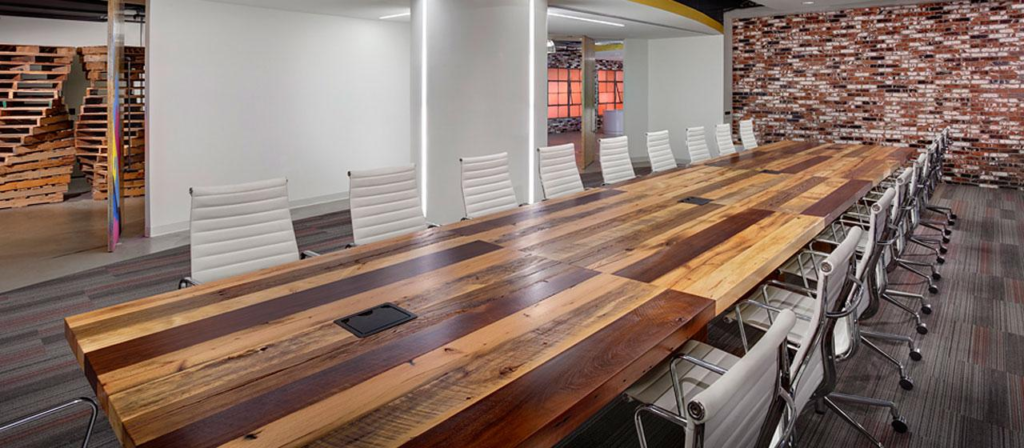 A modern conference room with a long table crafted from sustainable wood and white office chairs. The room features a brick accent wall, neutral-colored carpet, and soft lighting. The space appears open and contemporary.