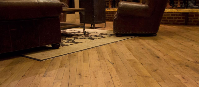 A warmly lit living room showcases leather chairs, a decorative rug, and a wooden coffee table with metal legs on distressed hardwood flooring. The brick walls enhance the cozy rustic look, creating an inviting atmosphere.
