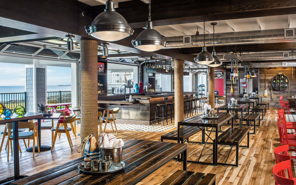 The restaurant boasts a spacious interior with stunning wood floors and rustic decor. Long tables with benches line the right, while a bar with industrial lighting is on the left. Large windows offer an ocean view, and various seating options are available throughout.