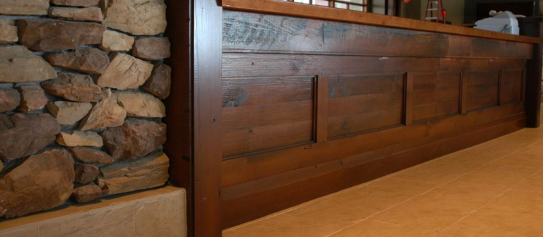 A wooden bar made from reclaimed wood with a polished surface features vertical paneling. To the left, a wall constructed of stacked rough rocks contrasts the wood. The floor is covered with square beige tiles. In the distance, a ladder is faintly visible.