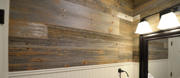 A bathroom with rustic reclaimed wood panel walls and a black-framed mirror exudes a cozy feeling. Three white light fixtures are mounted above the mirror, and a towel ring with a white towel hangs below. The lower half of the wall features classic white wainscoting.