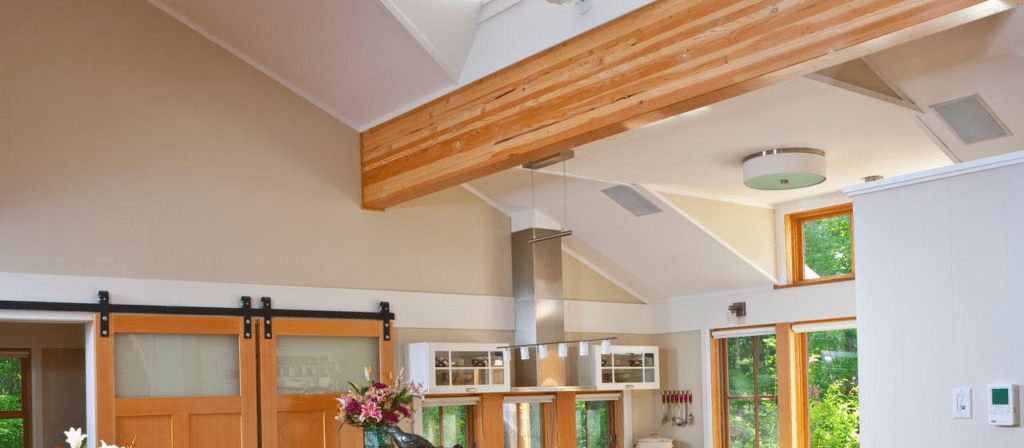 A modern kitchen with high ceilings and wooden beams boasts a central island adorned with a vase of flowers. A contemporary light fixture hangs above, while sliding doors made of sustainable wood complement the decor. Large windows invite natural light and offer views of greenery outside.
