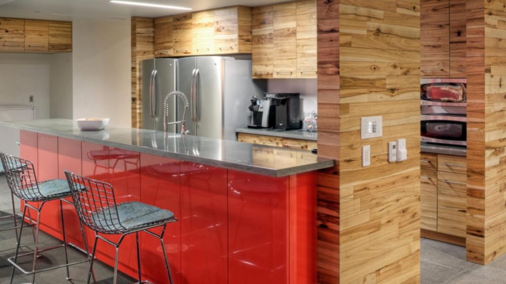 Modern kitchen with a red glossy island and two wire bar stools, doubling as an efficient work space design. Wooden cabinets and walls, stainless steel fridge, oven, and coffee maker are visible. The countertop features a curved faucet and a bowl. Soft lighting creates a cozy ambiance.