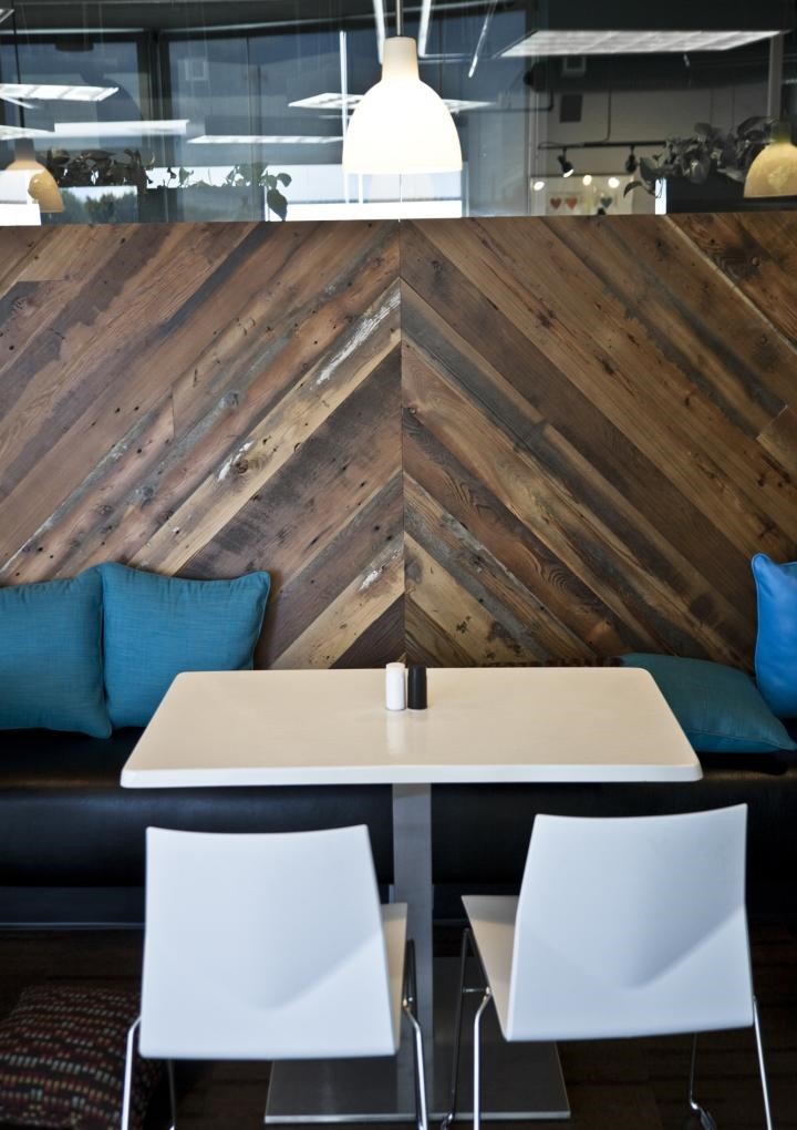 A modern café setting doubles as an inspiring work space design, with a white table and two chairs. The chevron-patterned wooden wall adds texture, while blue cushions on a black bench offer comfort. Pendant lights thoughtfully illuminate the area from above.