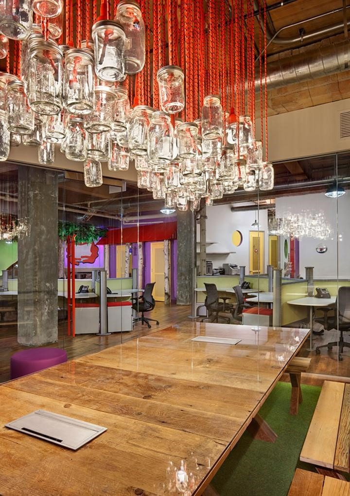 A modern office space boasts a well-thought-out work space design with a large wooden table and colorful chairs. The ceiling features a unique chandelier made of glass jars hanging from red strings, while open workstations with bright accents and exposed ductwork complete the vibrant atmosphere.
