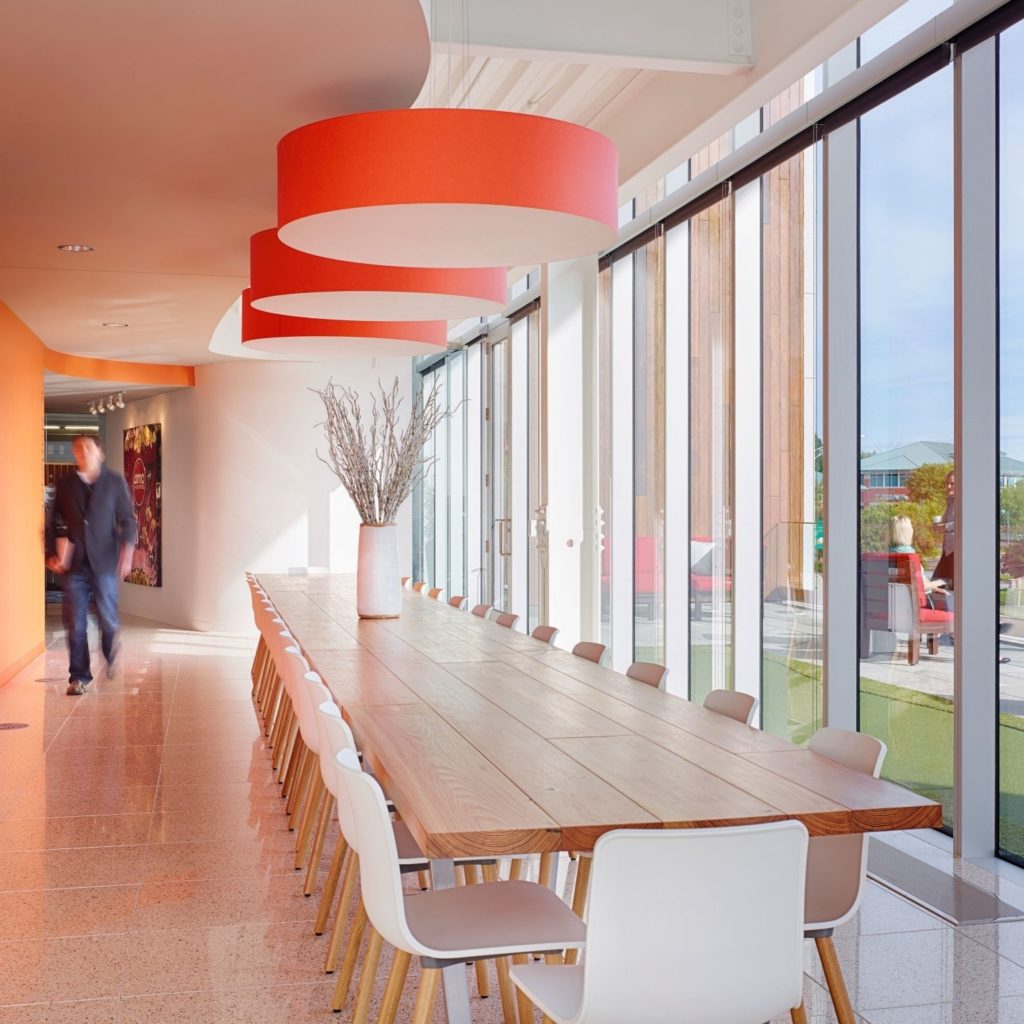 A modern work space design features a conference room with a long wooden table and white chairs. Large windows on the right let in natural light, while three large red circular lights hang from the ceiling. A person walks by an orange wall in the background, adding a touch of vibrancy.