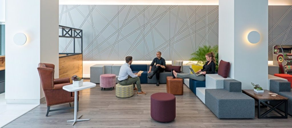 A modern office lounge with three people sitting on modular furniture showcases excellent work space design. The room features a geometric wall design, wooden flooring, a small side table with plants, and ambient lighting. A pinball machine is visible in the background.