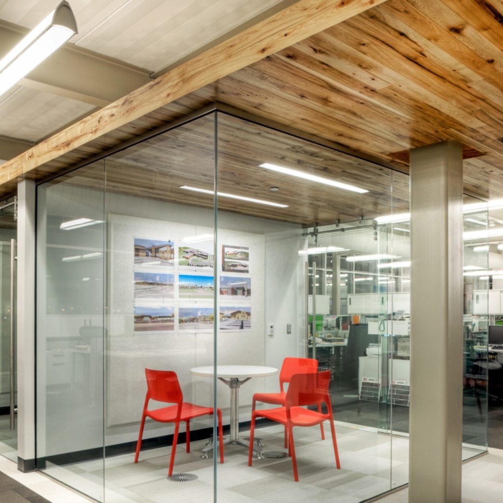 A modern work space design features glass walls, a small table with two red chairs, and a wooden ceiling. The gray carpeted floor complements several photos adorning one wall. This area is well-lit with overhead lights, offering a perfect blend of style and function.