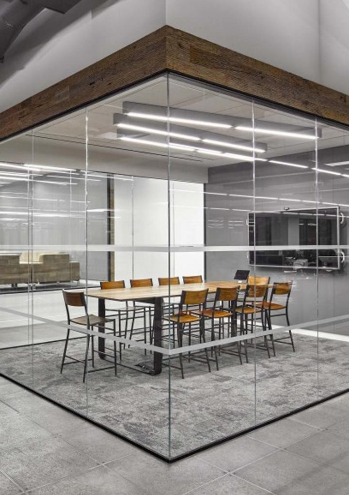 The modern work space design features a glass-walled conference room with a long white table surrounded by wooden chairs. It boasts gray carpet flooring, fluorescent lighting, and visible ceiling fixtures. Beyond the glass walls, the surrounding hallway adds an open feel to the area.