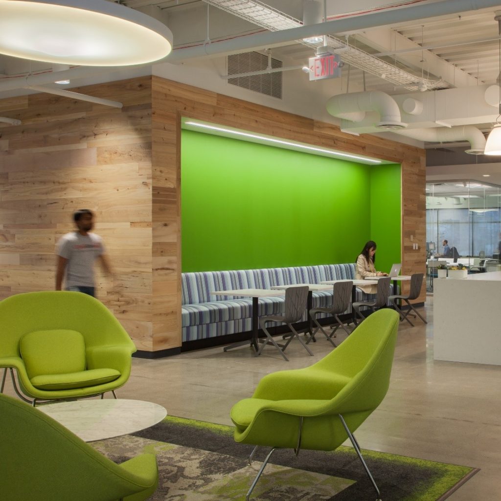 Modern office space with green accents, including chairs and a feature wall. One person sits at a long cushioned bench working on a laptop as another walks past. The area, epitomizing contemporary work space design, features wood paneling, overhead lighting, and an open layout.