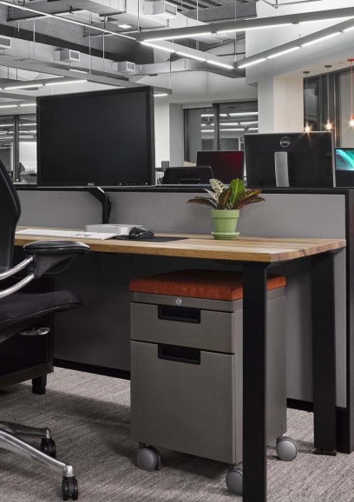 A modern office workspace boasts a sleek desk and ergonomic chair. This thoughtfully crafted work space design includes a computer monitor, keyboard, and a potted plant for greenery. Gray dividers, contemporary lighting, and a mobile gray filing cabinet complete the setup.