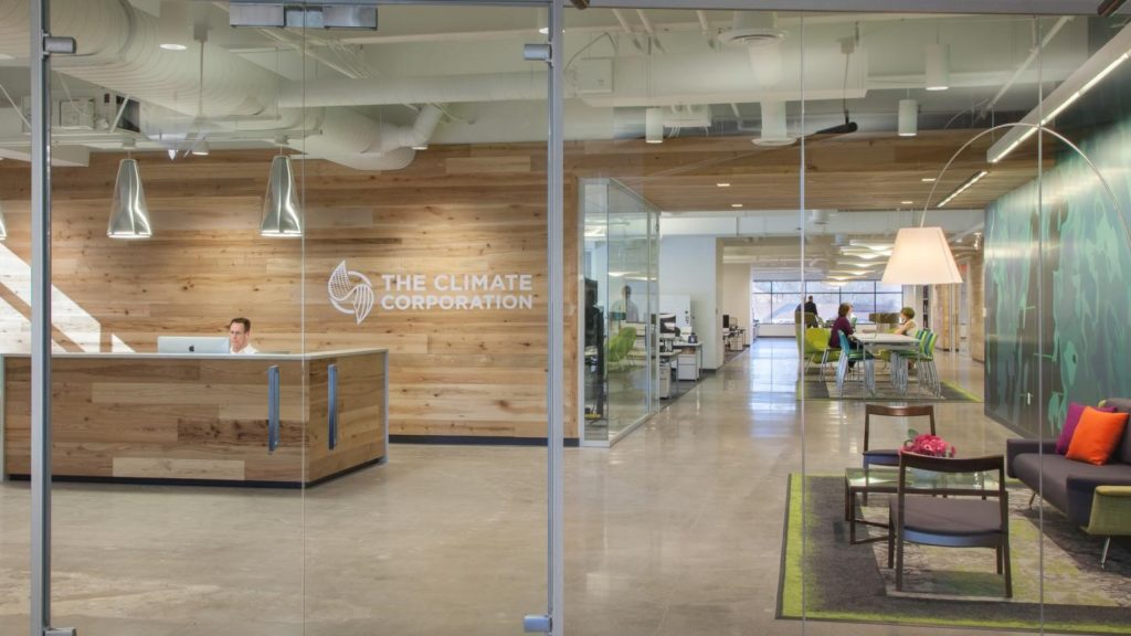 Modern office interior featuring innovative work space design with glass doors. On the left, a reception desk with a person seated beneath "The Climate Corporation" logo. On the right, a stylish lounge area offers chairs and a table, while the background reveals a corridor bustling with people at tables.