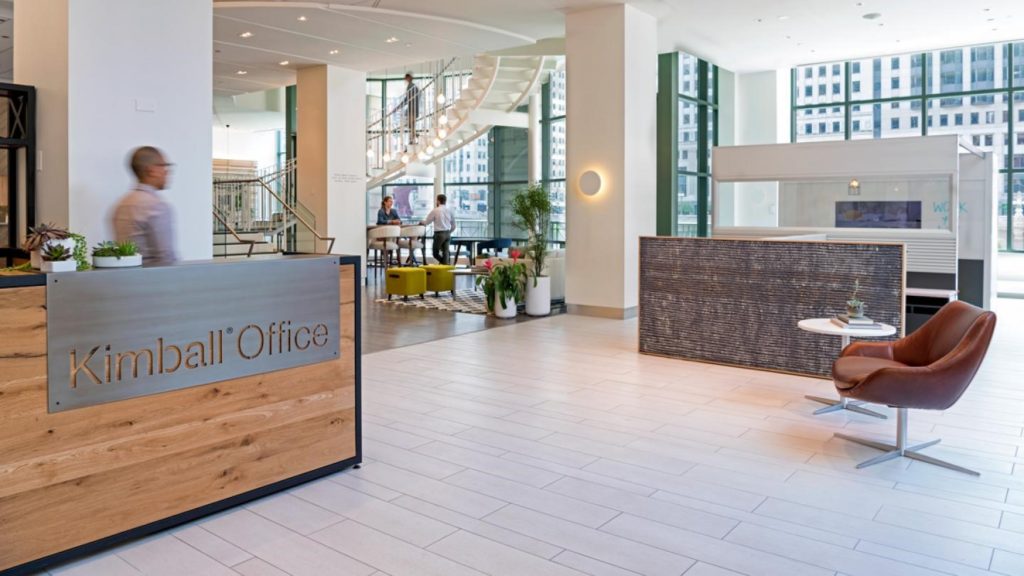 The modern office lobby showcases Kimball Office signage, featuring sleek work space design with wood and white decor. A hanging light fixture and spiral staircase complement the setting. Brown chairs and a gray reception area invite visitors, while large windows offer an urban view as people bustle in the background.