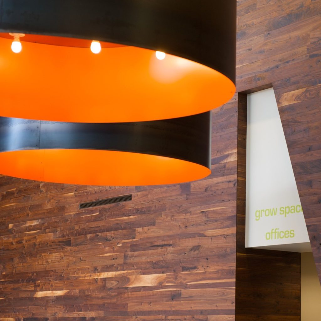 Large black cylindrical light fixtures with orange interiors hang against a backdrop of wooden panel walls, embodying modern work space design. To the right, a sign through a doorway reads "grow space offices.