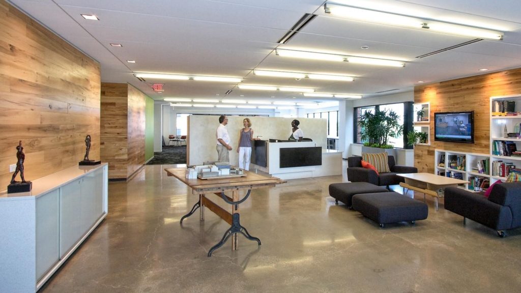 The modern office lobby showcases a sleek work space design with wooden accents, a polished concrete floor, and a welcoming reception desk. Three people are conversing by the desk, while comfortable seating with couches and book-filled shelves is on the right. Bright lighting and large windows illuminate the area.