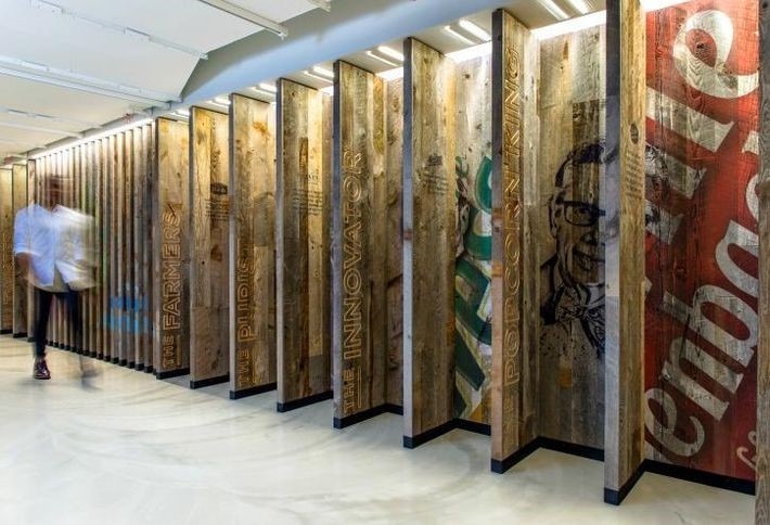 A person strolls past a series of vertical wooden panels adorned with colorful text and images. This innovative workspace design features panels that form a dynamic zigzag pattern along the corridor, enhanced by modern lighting and a polished floor.