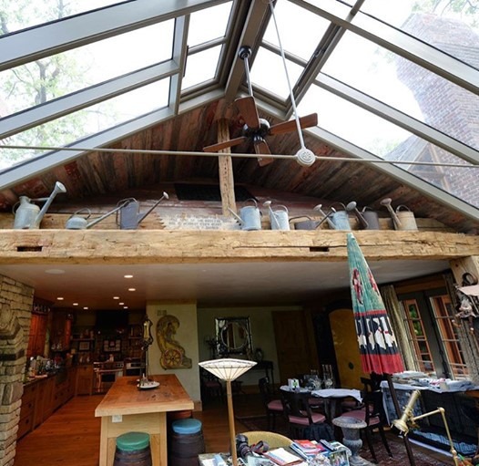 Interior design ideas shine through a skylight ceiling, rustic wooden decor, and various watering cans artfully displayed on a shelf. Below, a cozy room features a wooden table, umbrella, and assorted decor. Natural light fills the space, highlighting its eclectic style beautifully.