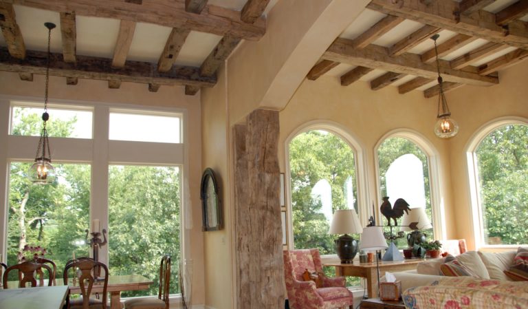 A cozy living and dining area showcases interior design ideas, with large arched windows offering lush greenery views. Exposed wooden beams adorn the ceiling, while the room features chairs, a sofa, a dining table, and stylish pendant lights.