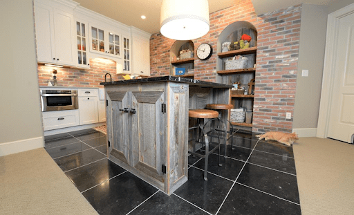A rustic kitchen features barnwood cabinets and a wooden island, complemented by two metal stools and black tile flooring. Exposed brick walls boast built-in shelves with decor. A large white pendant light hangs overhead while a small cat sleeps on the floor in the corner, making it feel like home.