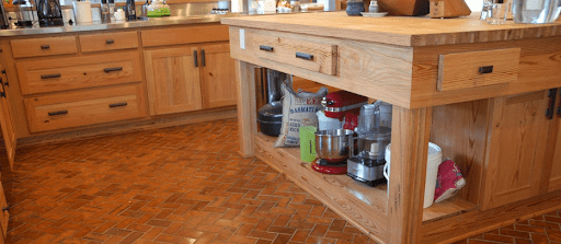 A charming kitchen features barnwood cabinets and brick-patterned flooring. The island, with open shelving, showcases a red stand mixer and a food processor. Countertops in this cozy home are delightfully cluttered with various essentials.
