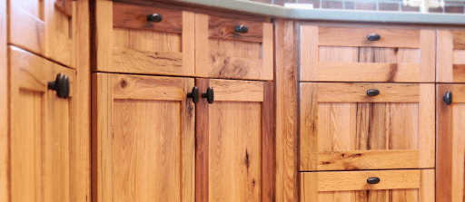 Close-up of barnwood kitchen cabinets with black handles, lending a rustic charm to your home. The cabinets boast a natural wood grain and warm tone, complemented by a countertop peeking above and a tiled backsplash in the background. Consider this blend for an inviting kitchen atmosphere.