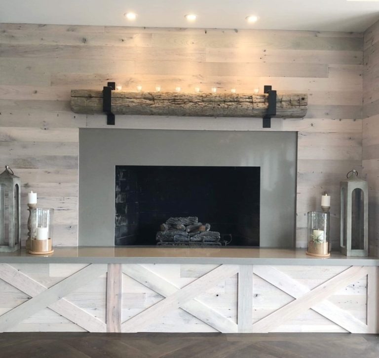 A modern fireplace with a rustic wooden mantel, decorated with lit candles, stands against a wall of whitewashed wood paneling. Two tall candle lanterns flank the hearth, seamlessly blending contemporary and rustic elements for a warm, inviting ambiance.