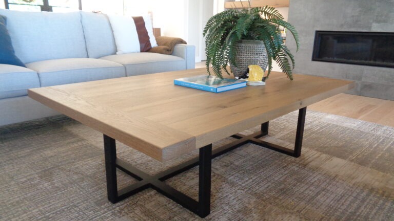 A modern living room showcases interior design trends with a light wooden coffee table featuring black metal legs. Adorning it are a book, a yellow decorative object, and a fern plant in a gray pot. The background is completed by a light gray sofa and fireplace.