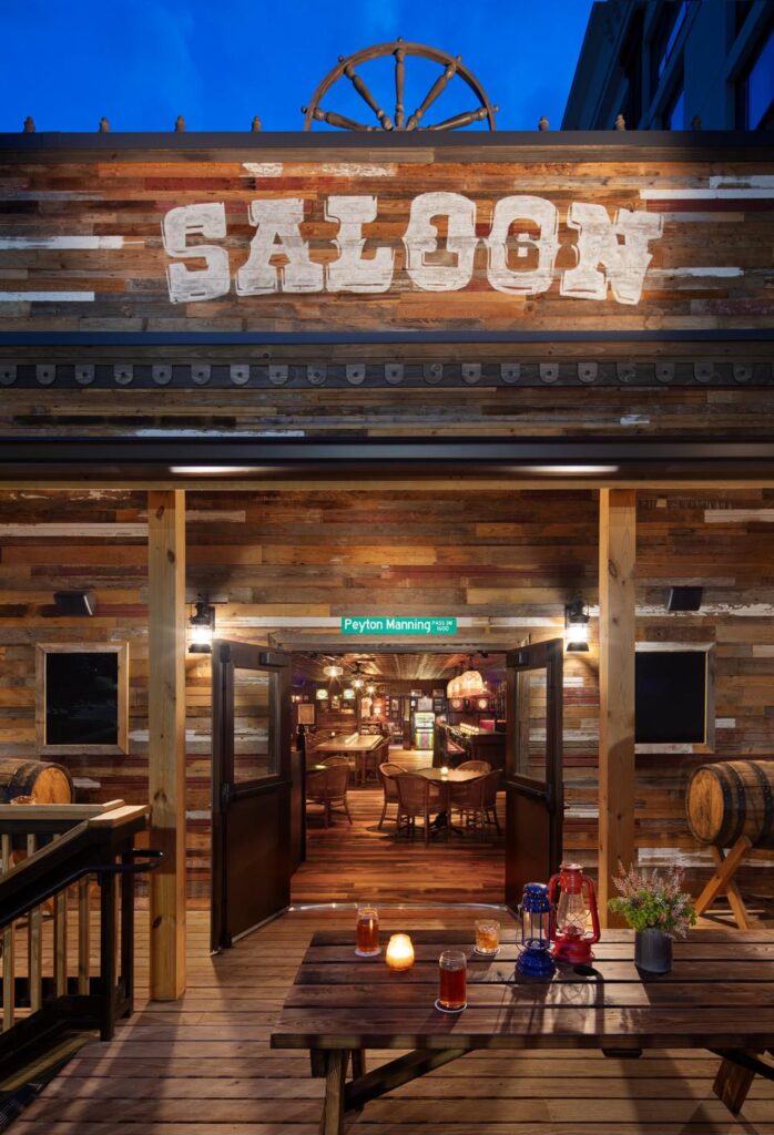A rustic saloon entrance made of barnwood siding, featuring a large "Saloon" sign above. The open doors reveal tables, chairs, and a warm, inviting interior. Barrels flank the entrance, and drinks are placed on the outdoor wooden deck.
