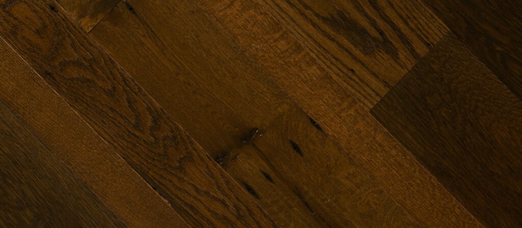 Close-up of dark wide plank floors with a rich, textured grain pattern. The planks vary slightly in tone, adding depth and warmth. Subtle variations in shading emphasize the natural wood finish and knots.