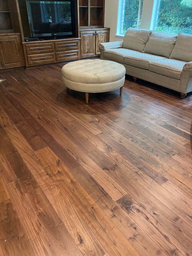 A living room adorned with walnut flooring, a beige couch by large windows, an upholstered round ottoman in the center, and a wooden entertainment unit with a TV.