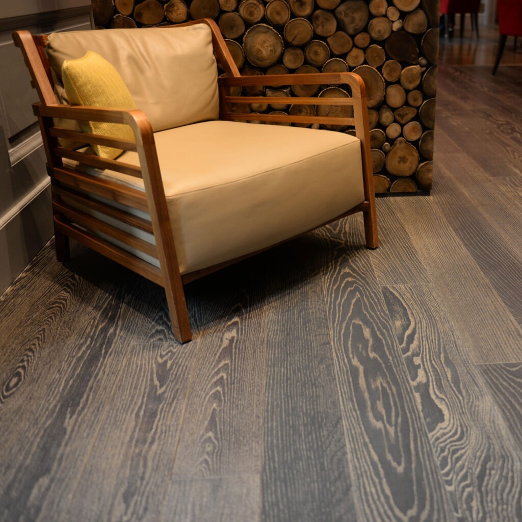 A tan cushioned armchair with a yellow pillow sits on elegant European White Oak flooring. Behind the chair, a decorative wall made of stacked circular wood logs adds rustic charm.