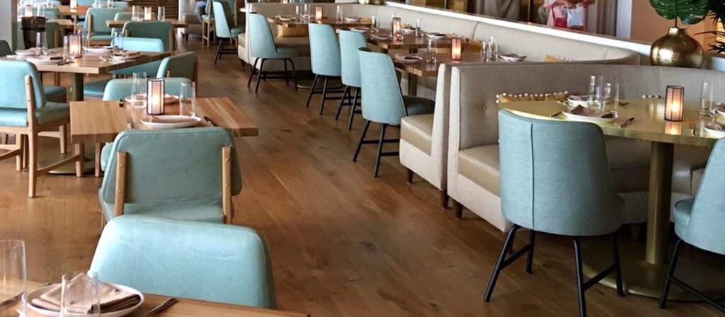 A stylish restaurant interior features light blue chairs and beige booths around wooden tables set with plates and glasses. The cozy atmosphere is enhanced by soft lighting and decorative accents on European Oak Flooring, creating an inviting ambiance on a wooden floor.