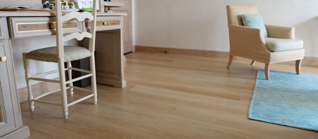 A room featuring European White Oak flooring showcases a wooden desk and chair on the left, complemented by a beige armchair with a light blue cushion on the right. A light blue rug graces the elegant engineered flooring beneath.