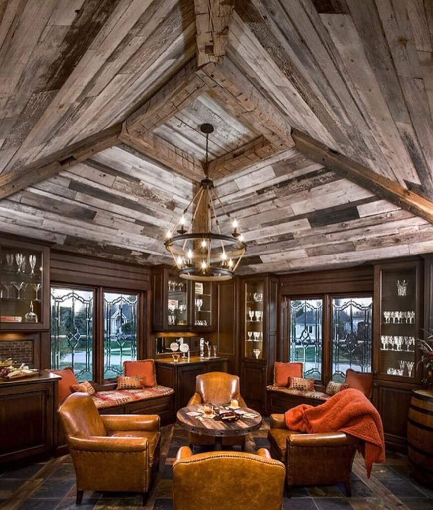 A cozy, rustic room with a wooden vaulted ceiling and wood wall paneling hosts a central chandelier. Leather chairs surround a round table, and bookshelves adorned with glassware line the walls. Orange throws add warmth to the seating area while large windows offer an outdoor view.
