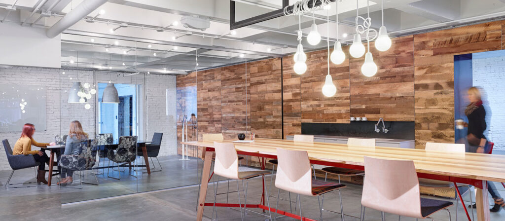 Modern office space featuring wood wall paneling, a long wooden table with barstools, and hanging light fixtures. Two people sit at a small table near a glass wall with a brick backdrop, while another person walks by on the sleek concrete floors.