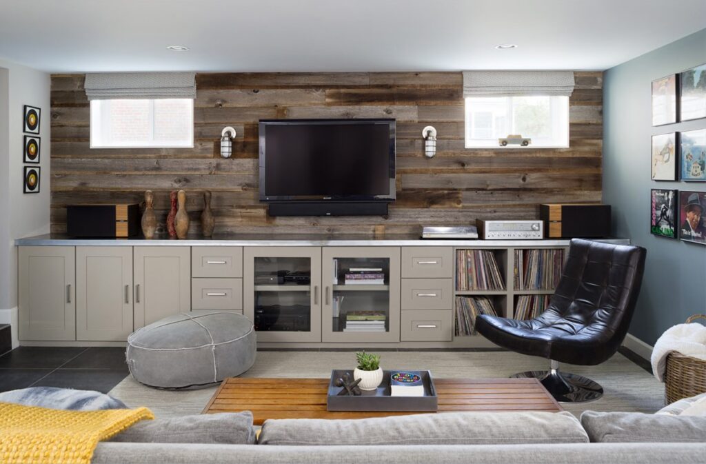 A modern living room features a TV mounted on rustic wood wall paneling. Below, cabinets house vinyl records and a stereo, while a black leather chair sits to the right and a gray ottoman to the left. Decor is completed with small sculptures and framed art.
