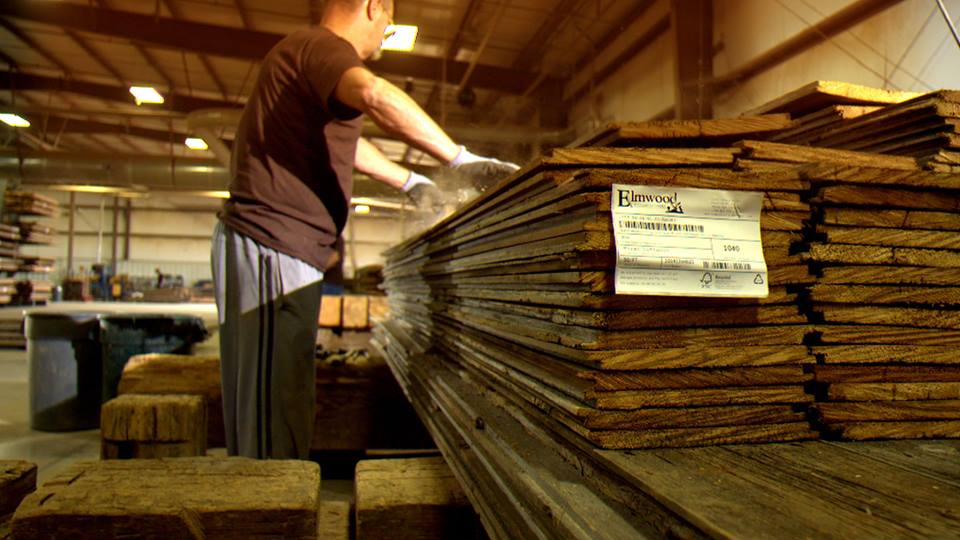 A person wearing a brown shirt and gloves efficiently stacks wooden planks labeled for green building projects in a spacious, well-lit warehouse with a high ceiling.