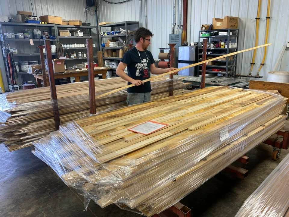 A person in a black T-shirt and glasses inspects a stack of wrapped, reclaimed wooden planks with a long stick in a workshop. Shelves with various supplies line the walls in the background.