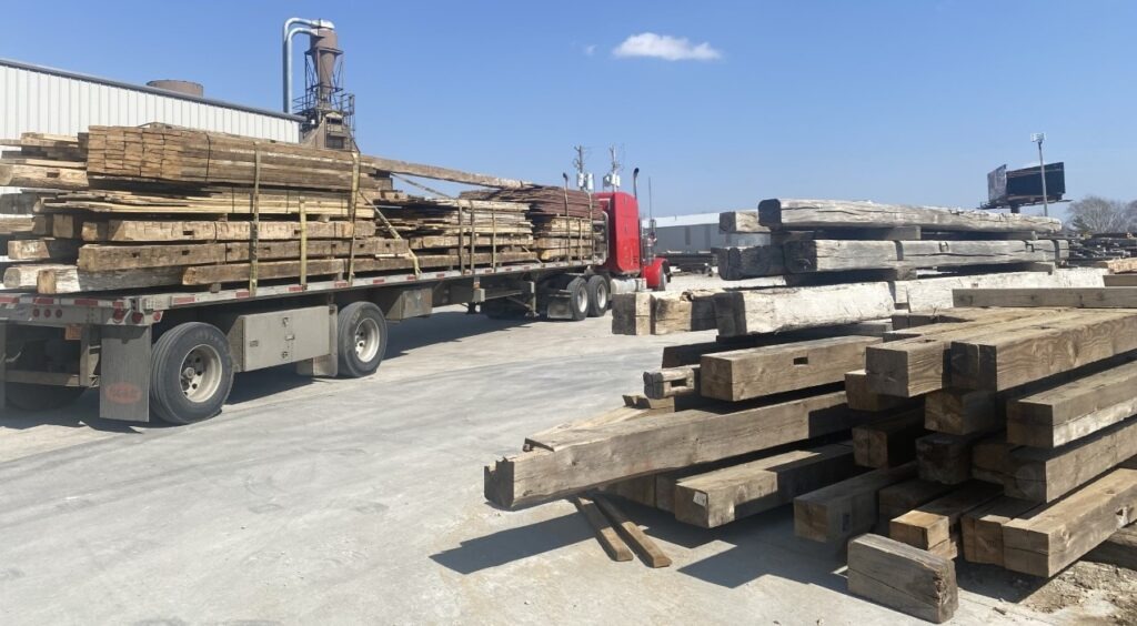 A red semi-truck carries large stacks of reclaimed wooden beams on a flatbed trailer, parked in an industrial area under a clear blue sky. More wooden beams are piled on the ground nearby. A building and machinery are visible in the background.