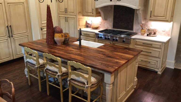 Rustic wood kitchen design with a central wooden island featuring a farmhouse sink and three woven-seat stools. The space is equipped with beige cabinetry, a gas stove under a white hood, and stone countertops. Dark wooden floors beautifully complement the warm tones.