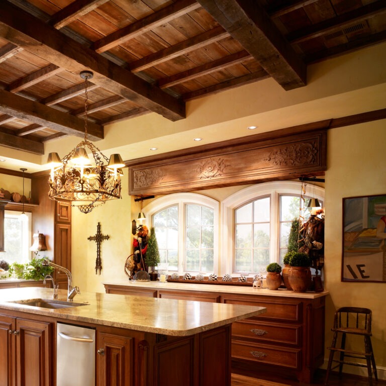 A rustic kitchen features reclaimed wood cabinets, an island with a stone countertop, a decorative chandelier, and a cross on the wall. There are arched windows adorned with plants, letting in natural light, and exposed wooden beams on the ceiling.