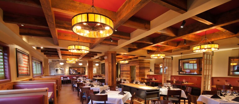 A warm, inviting restaurant interior features box beams and amber chandeliers. Tables are neatly set with white tablecloths and dark chairs. Red cushioned booths line the walls, while soft lighting creates a cozy atmosphere.