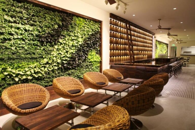 A modern café interior with round wicker chairs and wooden tables crafted from eco-friendly materials. A lush vertical garden decorates the wall on the left, while the right side features a bar with rich wood accents. The warmly lit space boasts a tiled floor for a cozy ambiance.