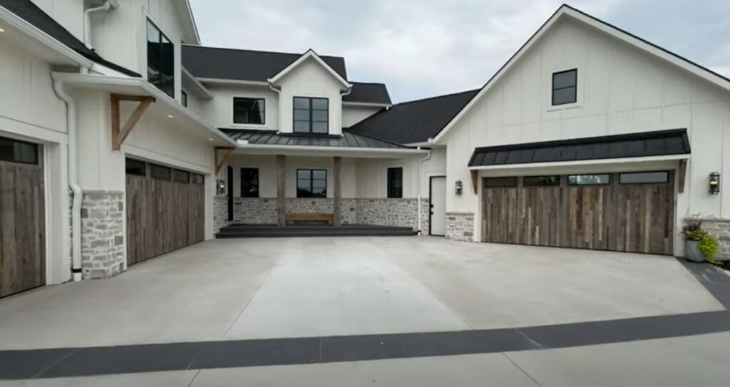 The modern suburban house at Stone Springs Ranch features white siding, stone accents, and a sleek black roof. It includes three wooden garage doors and a covered entrance with a bench. The spacious driveway leads up to the entrance under an overcast sky.